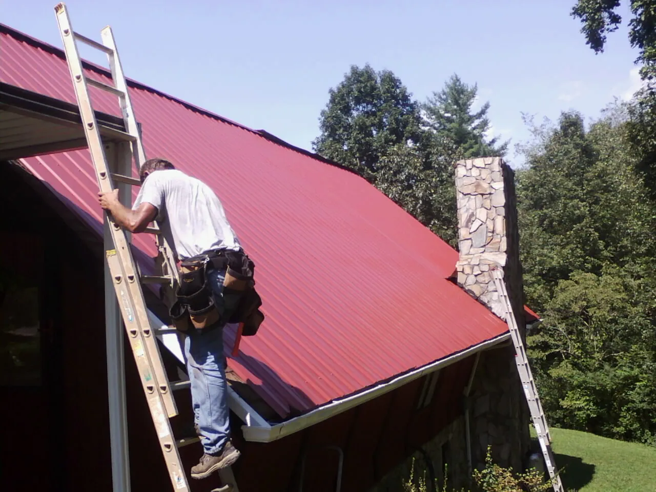 Roofer on a ladder beside a red metal roof and stone chimney — Soffit & Fascia Repair by Roofing Brothers in Carmen